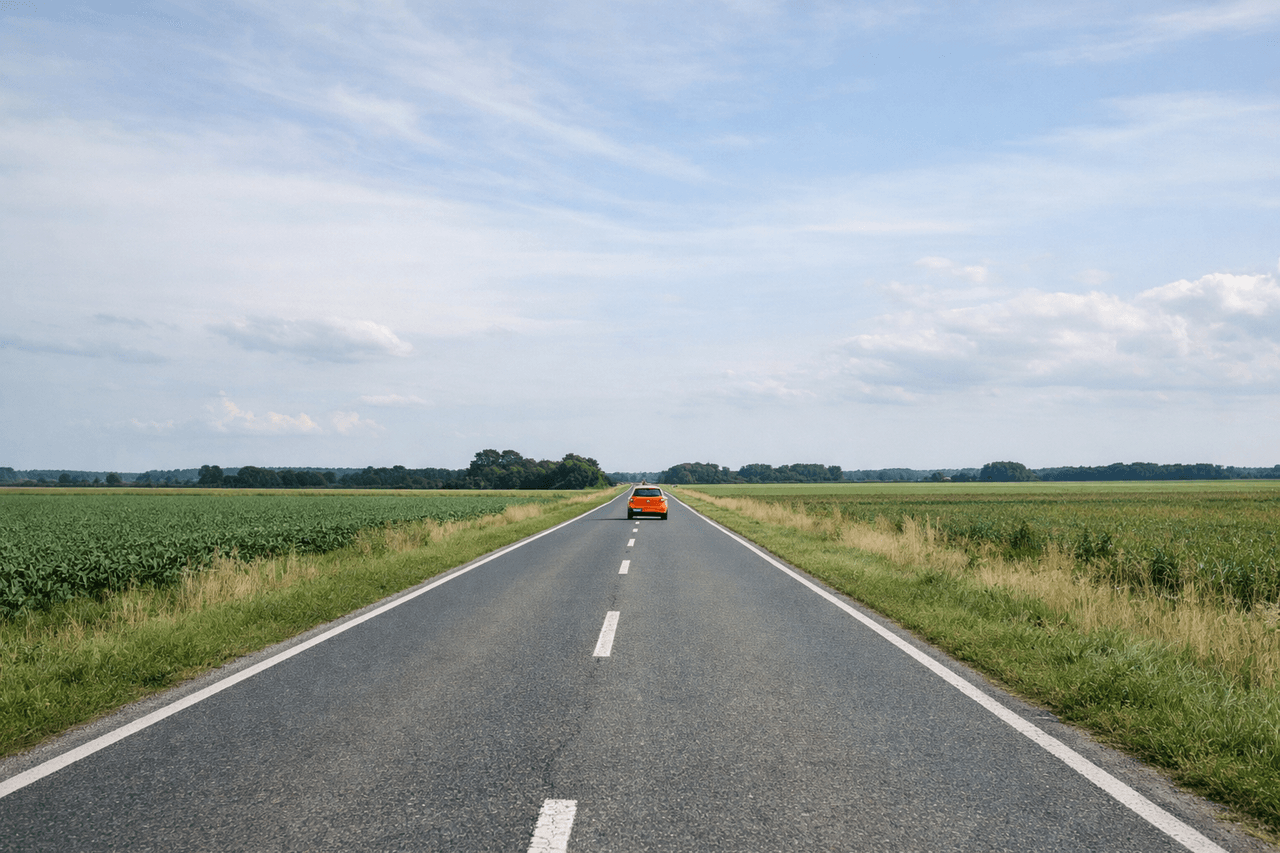Gerade Landstraße zwischen grünen Feldern unter weitem Himmel, in der Ferne ein oranges Auto